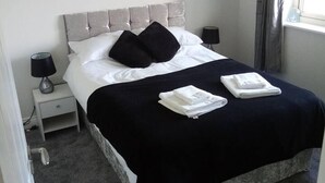 Un lit bien fait, avec une tête de lit grise, deux oreillers noirs et une couvre-lit noire. On y trouve deux tables de chevet avec des lampes, ainsi qu’une fenêtre avec des rideaux.