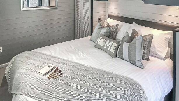 Un lit bien fait, recouvert d’une couvre-lit à motifs, avec des oreillers sur lesquels est brodé le mot « HOME », et une table de chevet sur laquelle se trouvent un livre et un petit objet.