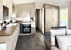 A compact kitchen with modern appliances, a dining area with a sofa, and a view through an open door. - Valley View 2 Bed Hot Tub Holiday Caravan Cawsand (St Ann&rsquo;s Chapel, Callington)