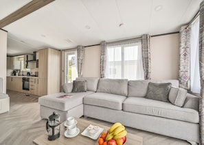 A modern living room with a grey sectional sofa, a coffee table with fruits and a teapot, and a kitchen in the background.