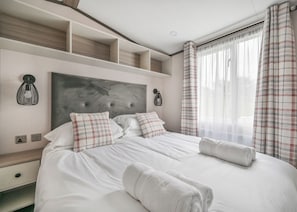 A double bed with white linens, plaid pillows, and a headboard with a textured design. There are two wall-mounted light fixtures and a window with plaid curtains.