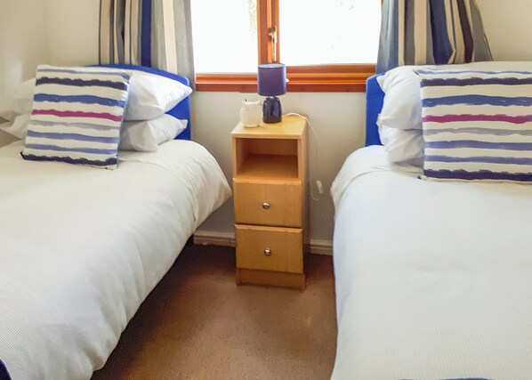 Una habitación con dos camas, cada una con almohadas a rayas, una mesita de noche con lámpara y una ventana con cortinas.