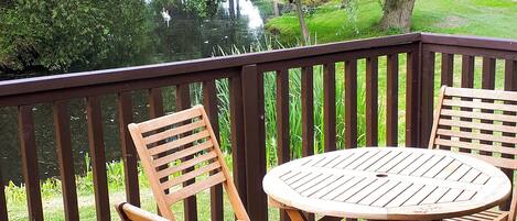 A wooden deck with a round table and two chairs, overlooking a pond and greenery.