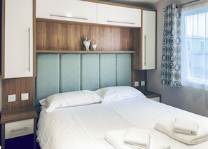 A bedroom with a bed, two nightstands, a wooden headboard, a window with patterned curtains, and a wall-mounted cabinet. - Merrick Platinum Caravan (Glenluce, Newton Stewart)