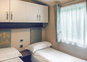 A bedroom with two single beds, a wardrobe, a window with sheer curtains, and a decorative headboard.