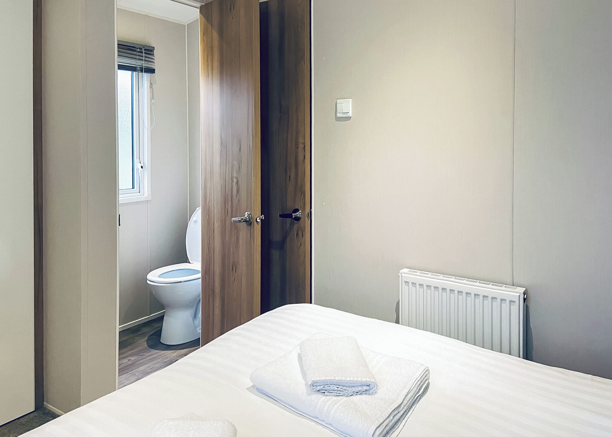 Une chambre à coucher avec un lit, un radiateur blanc, une porte en bois et des toilettes visibles à travers une porte entrouverte.