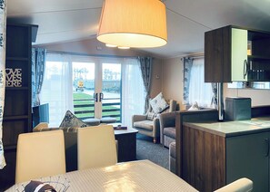 A compact living space with a dining table, two sofas, a kitchen area, and large windows offering a view of the outdoors.