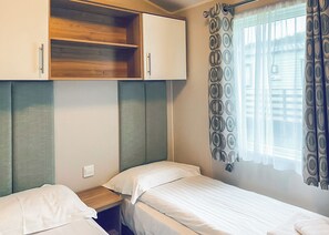 Une chambre avec deux lits simples, une étagère en bois et une fenêtre agrémentée de rideaux à motifs.