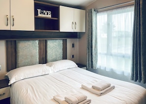 A hotel room with a bed, a wardrobe, a shelf with a 'LOVE' sign, and a window with curtains. - Merrick Platinum Caravan (Glenluce, Newton Stewart)