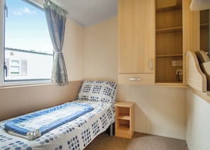 Ein Einzelbett mit einem Punkte-Bettbezug, ein kleiner Nachttisch aus Holz, eine eingebautes Holzschränkchen mit einer Schublade und ein Fenster mit Vorhängen.