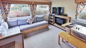 A cozy living room with a sofa, TV, wooden entertainment unit, and a dining table. - The Gold Plus 2 caravan is 12ft wide with one double and one twin. (Clarach Bay, Nr Aberystwyth)