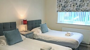 A hotel room with two beds, a nightstand, a lamp, and a window with blinds.