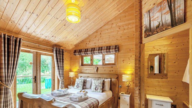 A wooden cabin bedroom with a bed, bedside lamps, a nightstand, and a window overlooking greenery. - 2 bedroom accommodation in Ellesmere (Ellesmere)