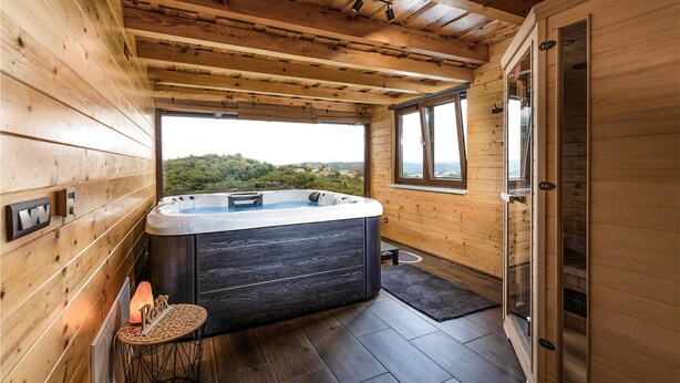 A wooden cabin with a hot tub, a small table with candles, and a view of the landscape.