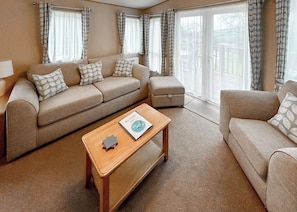 A living room with a sofa, ottoman, and coffee table, large windows with curtains, and a view of the outdoors. - Country Holiday Home Platinum Four Spa (Tintagel)