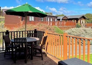 Um deck de madeira com mesa e cadeiras, cercado por uma vedação de madeira e um guarda-sol verde.