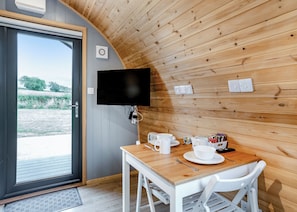 Une pièce avec un plafond en bois, un téléviseur fixé au mur, une table avec des chaises blanches et une porte donnant sur l’extérieur.