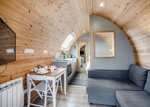 A cozy, compact room with a wooden ceiling, a dining area with a table and chairs, a grey sofa, and a kitchen visible through an archway.