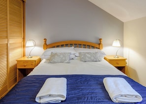 Ein ordentlich bezogenes Bett mit blauer Tagesdecke, zwei weiße Handtücher und zwei Nachttischlampen.