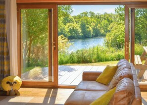 A room with a sliding glass door leading to a deck overlooking a lake, a leather sofa with cushions, and a small decorative sheep figure. - Perfectly positioned with panoramic views of the lake. (Anglesey)