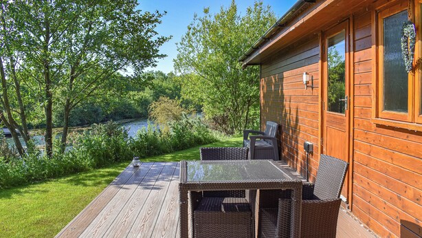 A wooden deck with a table and chairs, a view of a lake, and a cabin with windows. - Perfectly positioned with panoramic views of the lake. (Anglesey)
