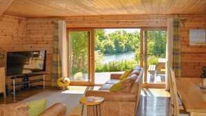 A wooden cabin interior with a sofa, TV, and dining area, overlooking a lake. - Perfectly positioned with panoramic views of the lake. (Anglesey)