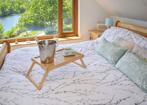 A bedroom with a bed, a bedside table with a lamp, a radio, and a view of a river and trees.