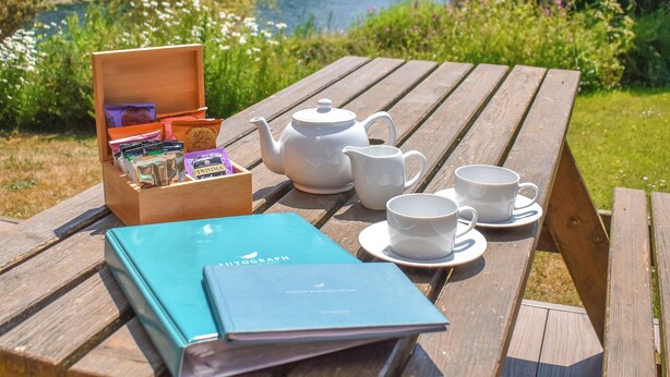 Une table de pique-nique en bois, avec une théière, des tasses, une boîte de collations et un livre.