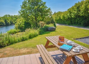 A wooden deck with a picnic table set up overlooking a river, surrounded by greenery and trees. - Perfectly positioned with panoramic views of the lake. (Anglesey)