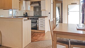 A compact kitchen with wooden cabinets, a central island, a stove, and a dining area with chairs. - Sunnyvale Willow 3 Pet Friendly (Saundersfoot)