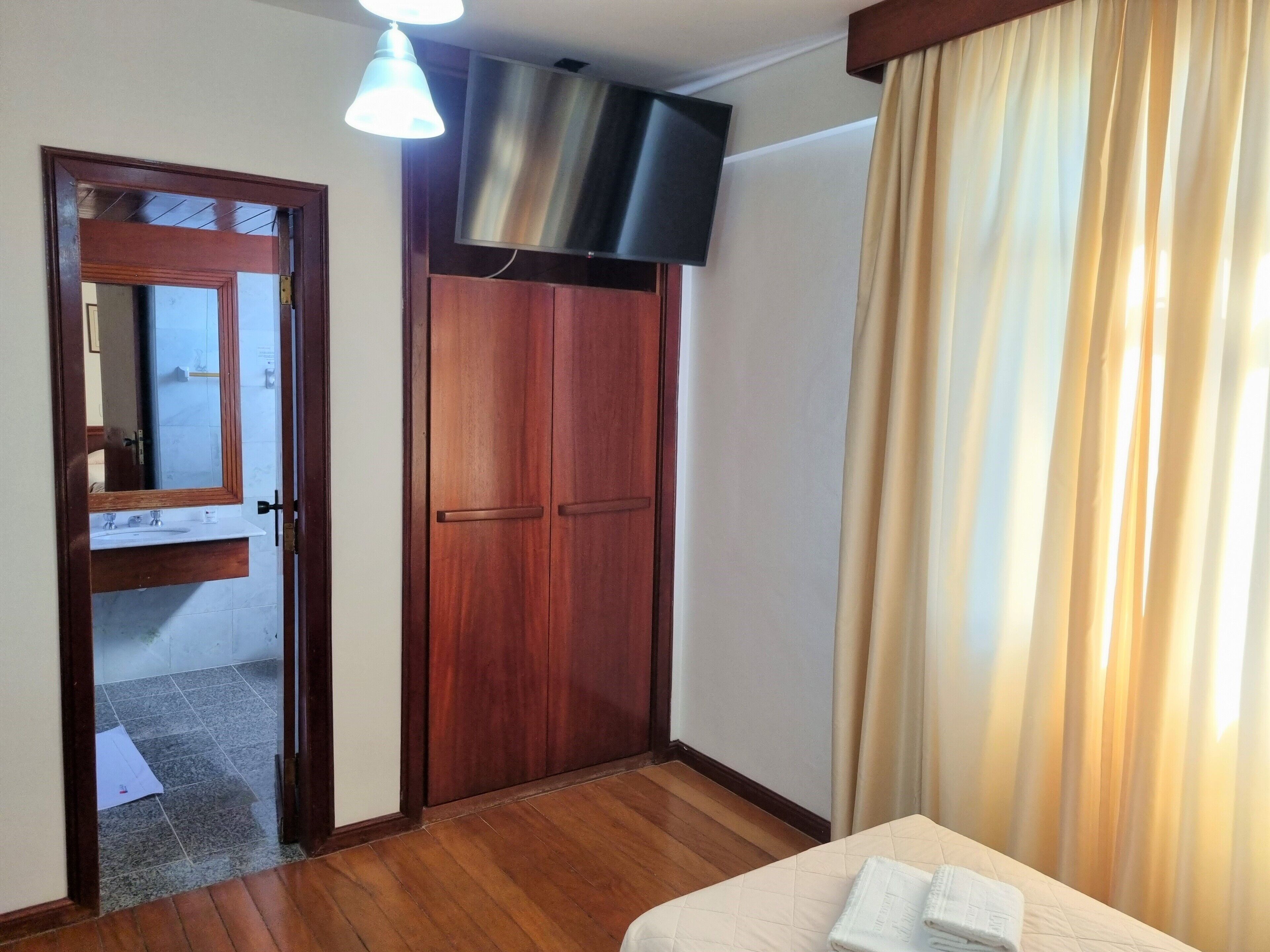 A hotel room with a wooden wardrobe, a television mounted above it, a bed with a white duvet, and a window with beige curtains.