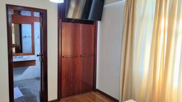 A hotel room with a wooden wardrobe, a television mounted above it, a bed with a white duvet, and a window with beige curtains.