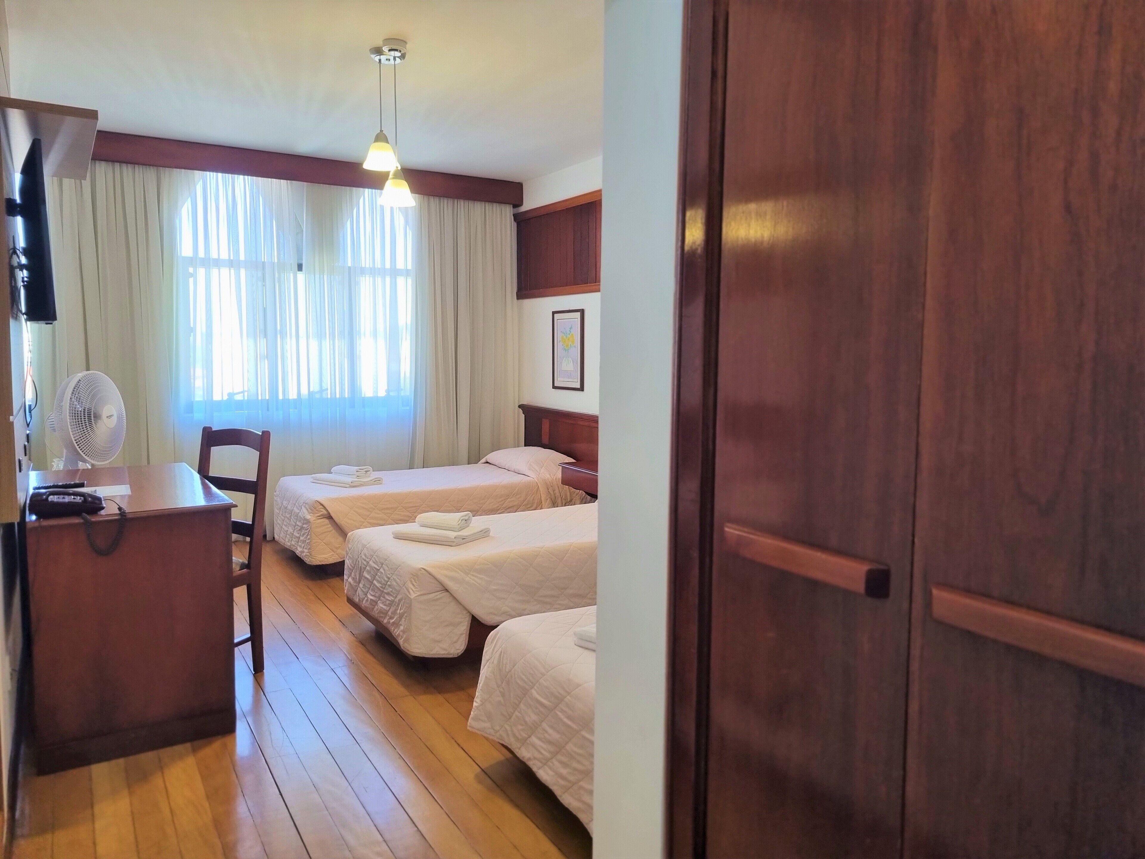 A hotel room with two beds, a desk with a telephone, a chair, a fan, and a wooden door.