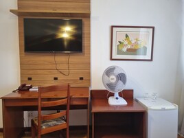 Un escritorio de madera con un televisor montado encima, una obra de arte floral enmarcada en la pared, un ventilador de pie y una mesita con un teléfono y un libro.