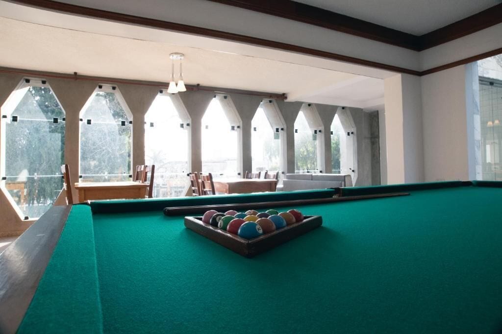 Una mesa de billar con superficie de fieltro verde, un juego de bolas de colores y un taco. Hay ventanas arqueadas con vistas al exterior.