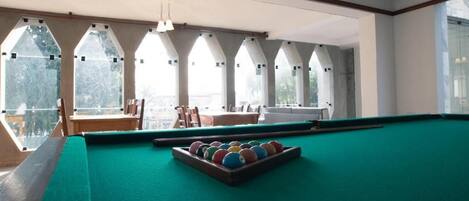 Una mesa de billar con superficie de fieltro verde, un juego de bolas de colores y un taco. Hay ventanas arqueadas con vistas al exterior.