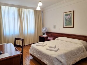 A hotel room with a bed, a bedside table, a chair, a desk, a window with curtains, a wall-mounted fan, and a framed picture. - Lucape Palace Hotel (Barbacena)