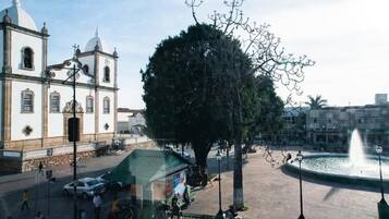 Uma praça com uma fonte, uma árvore grande e uma igreja com duas cúpulas.