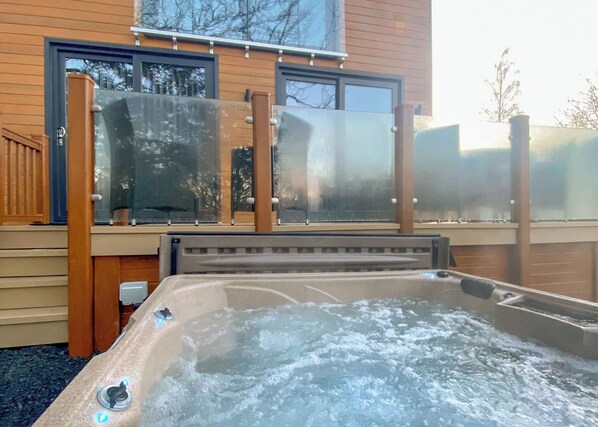 A hot tub on a wooden deck with glass railings, a window, and a view of trees. - 1 bedroom accommodation in Water Yeat, Nr Coniston (Water Yeat, Nr Coniston)