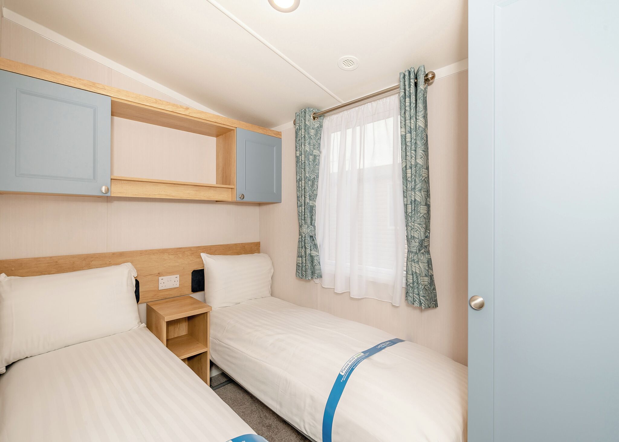 Una habitación pequeña y bien iluminada con dos camas, cabeceros de madera, un estante y una ventana con cortinas.