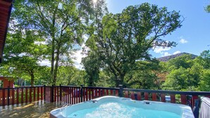 A hot tub on a deck with a wooden railing, surrounded by trees and a mountain in the background.
