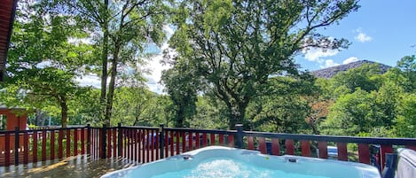 A hot tub on a deck with a wooden railing, surrounded by trees and a mountain in the background.