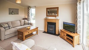 A living room with a sofa, coffee table, fireplace, TV stand, and a painting on the wall. - Contemporary lodge furnished to a very high standard. (Tunstall, Nr Hull)