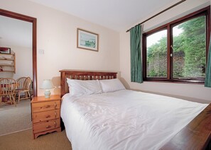 A bedroom with a wooden bed, a bedside table with a lamp, a chair, a framed picture on the wall, and a window with curtains.