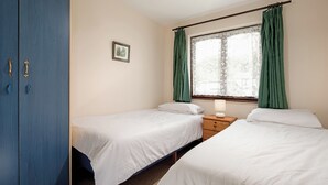 A room with two single beds, a wooden wardrobe, a small bedside table with a lamp, and a window with green curtains. - Silver Bungalow 2 (sleeps 4) (Penzance)