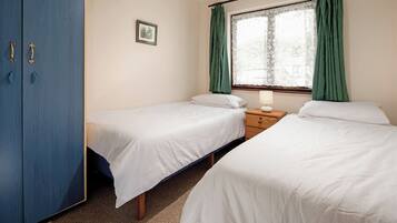 A room with two single beds, a wooden wardrobe, a small bedside table with a lamp, and a window with green curtains.