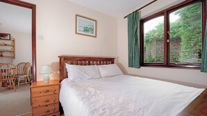 Une chambre avec un lit en bois, une table de chevet avec une lampe, une chaise, un tableau encadré au mur et une fenêtre avec des rideaux.