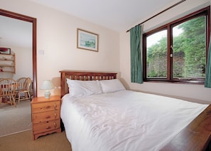 A bedroom with a wooden bed, a bedside table with a lamp, a chair, a framed picture on the wall, and a window with curtains.