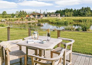 A wooden deck with a table and chairs, a candle holder, and a bottle of champagne. In the background, there is a pond, a wooden building, and a grassy area with trees. - A stunning luxury lodge, perfect for a couples retreat or family break. (West Tanfield)