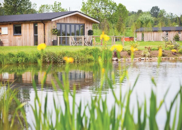A wooden building with a large window and a door, a small garden with a pond, and a fence. - A stunning luxury lodge, perfect for a couples retreat or family break. (West Tanfield)